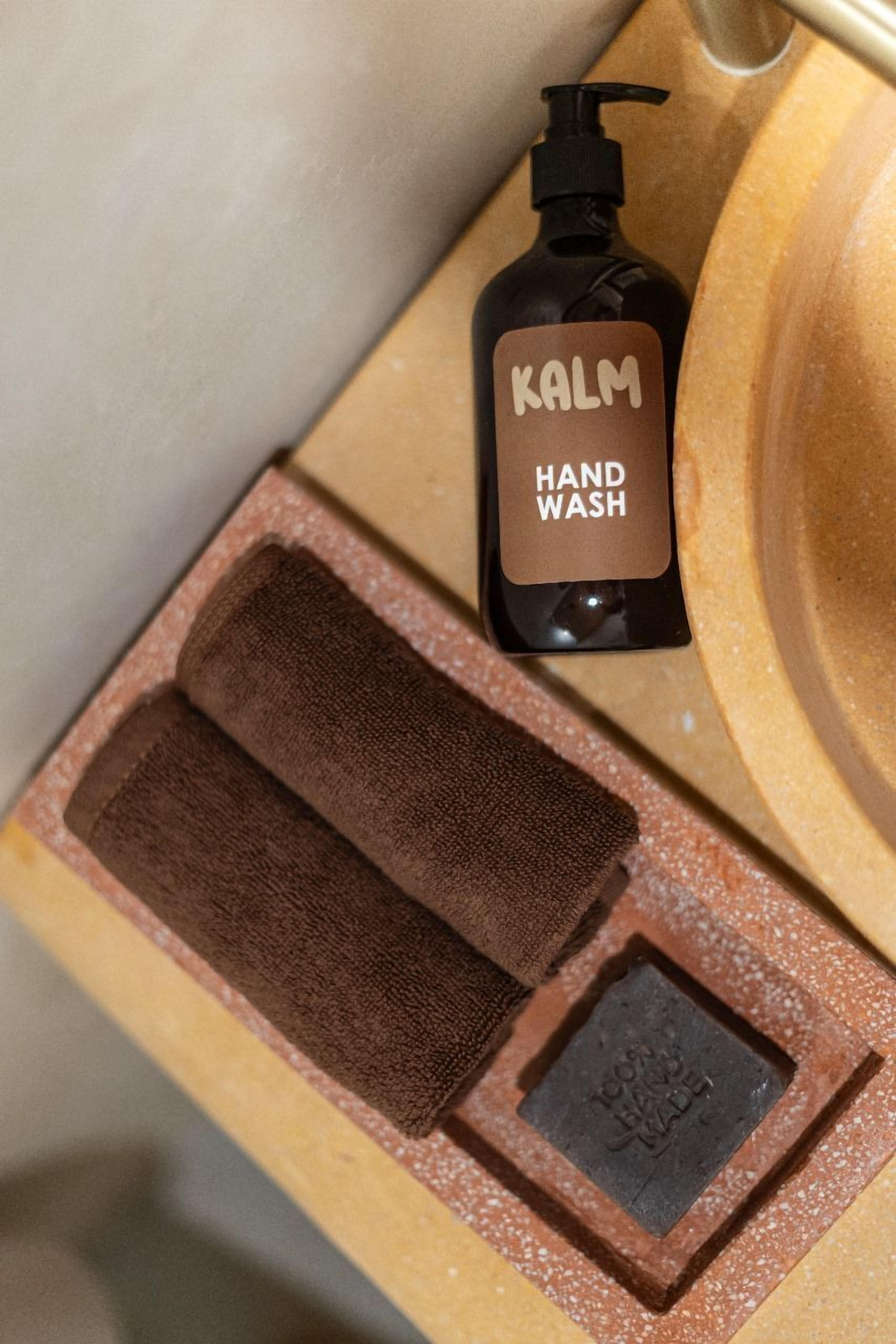A hand wash soap bottle labeled 'KALM HAND WASH' next to a wooden sink with a brown towel and a soap dish.