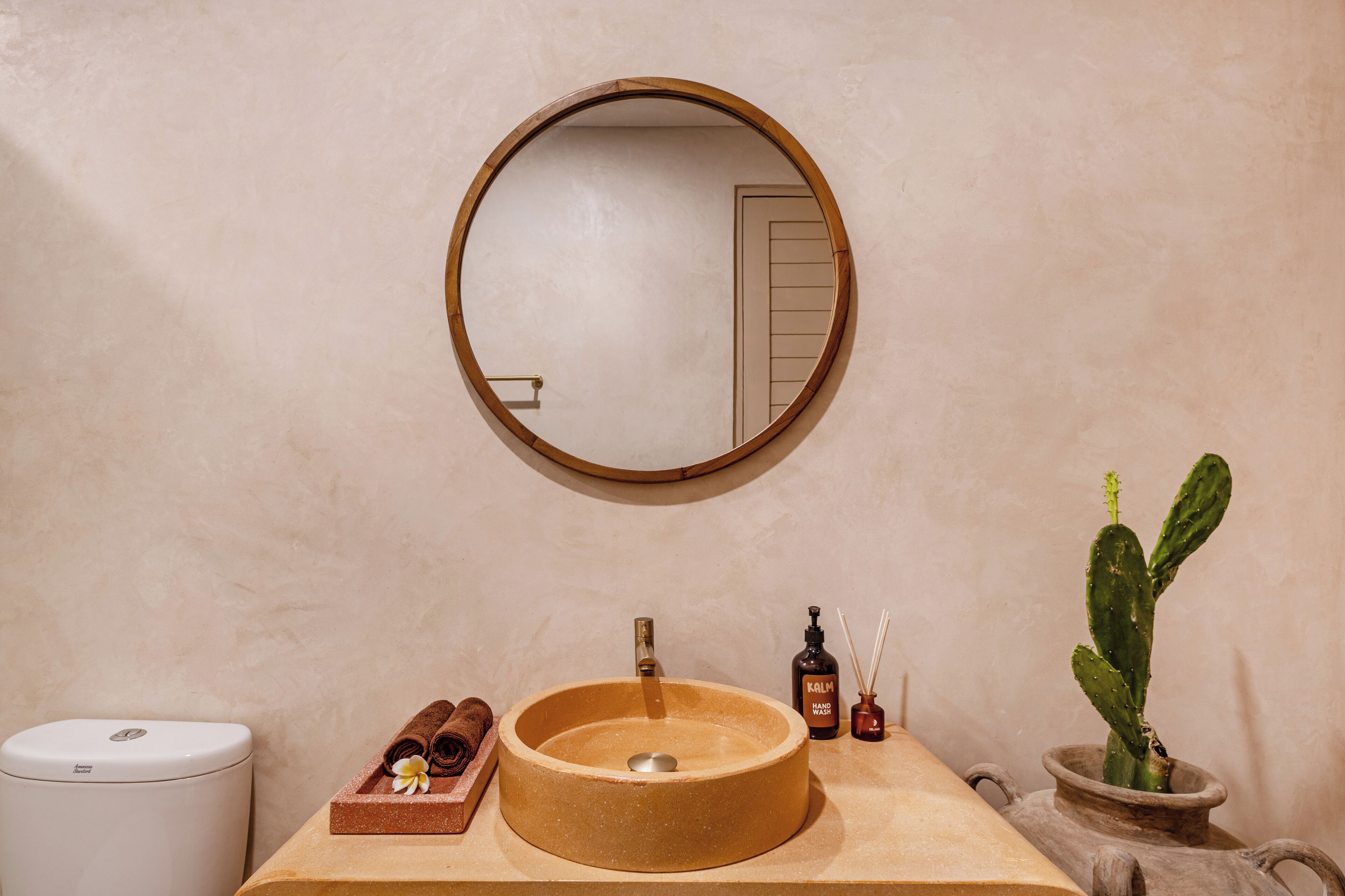 Een badkamer met een ronde spiegel, een wastafel, een toilet, een handdoek, een fles en een cactus.