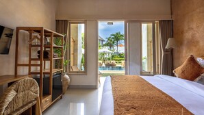 Une chambre d’hôtel avec un lit, un bureau en bois, une chaise, une armoire et une vue sur une piscine et des palmiers.