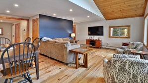 A spacious living room with a wooden ceiling, a large TV, and a dining area with a table and chairs. - Lakeside Lodge at Fern Lake: 3BR/1BA Waterfront w/ Dock (Leicester)