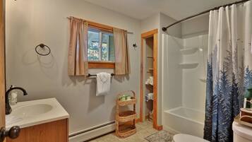 A bathroom with a sink, toilet, and shower. There is a window with curtains, a towel rack, and a wicker basket.