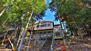 A cabin with a wooden deck and a staircase leading up to it, surrounded by trees. - Lakeside Lodge at Fern Lake: 3BR/1BA Waterfront w/ Dock (Leicester)