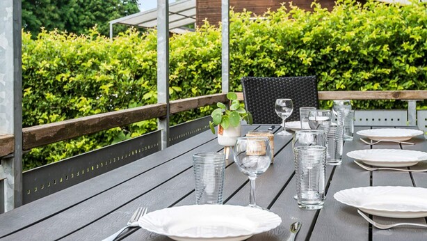 Un espace de restauration en plein air avec une table dressée pour quatre personnes, entouré de verdure et avec un bâtiment en arrière-plan.