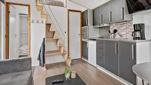 Une cuisine compacte avec des meubles gris, un four encastré et une machine à café. Un escalier en bois mène à une mezzanine.