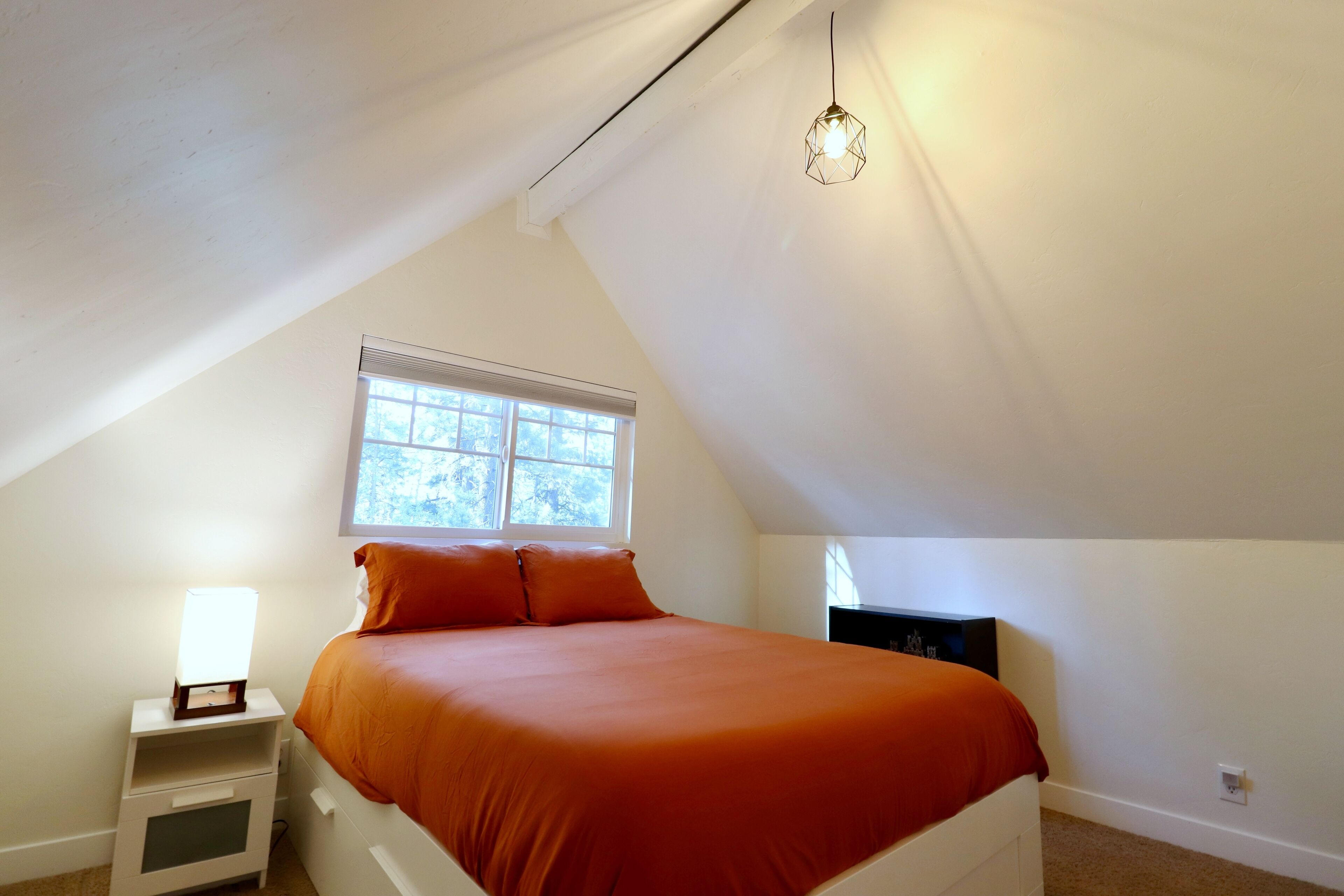 Un dormitorio con techo inclinado, una cama con ropa de cama naranja, una mesita de noche con lámpara y una ventana que da a vistas de árboles.