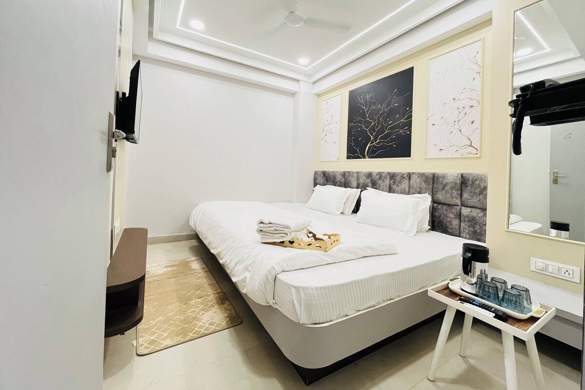 Une chambre d’hôtel moderne avec un lit soigneusement fait, une table de chevet avec une machine à café et des verres, et des œuvres d’art accrochées aux murs.