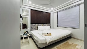 A modern bedroom with a large bed, a ceiling fan, a bedside table with a lamp, and a window with blinds. - Hotel Padmawati Palace (Ujjain)