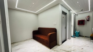 A modern room with a brown sofa, white marble flooring, recessed ceiling lighting, a red fire extinguisher, and a blue trash bin. - Hotel Padmawati Palace (Ujjain)