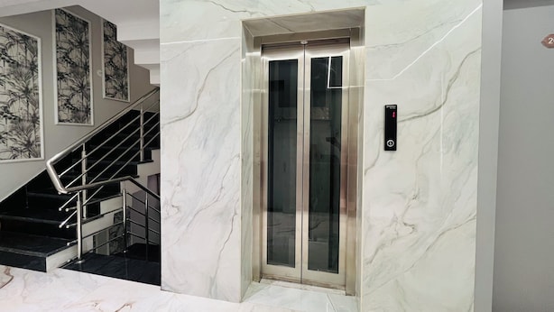A modern elevator with a marble wall and a glass door, a staircase with metal railings, and a wall with framed botanical prints. - Hotel Padmawati Palace (Ujjain)