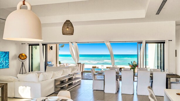 Un salón moderno con una amplia vista al océano, muebles blancos y una zona de comedor con sillas blancas.