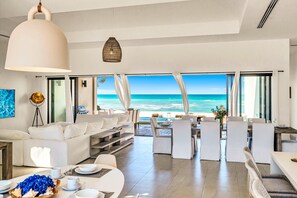 Un salon moderne offrant une vue imprenable sur l’océan, avec des meubles blancs et un coin repas doté de chaises blanches.