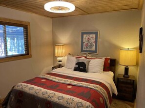 Une chambre à coucher comprenant un lit, deux lampes de chevet, une fenêtre avec des stores, un luminaire au plafond et une œuvre d’art florale encadrée accrochée au mur.