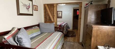 A small, cozy room with a bed, a wooden dresser, a television, and a door leading to another room.