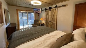 A bedroom with a bed, a TV mounted on the wall, a sliding barn door, and a window with a view of the outdoors. - Midtown Meadow Cabin (Ruidoso)