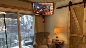A room with a sliding glass door, a cushioned armchair, a table lamp, and a television displaying a cityscape. - Midtown Meadow Cabin (Ruidoso)