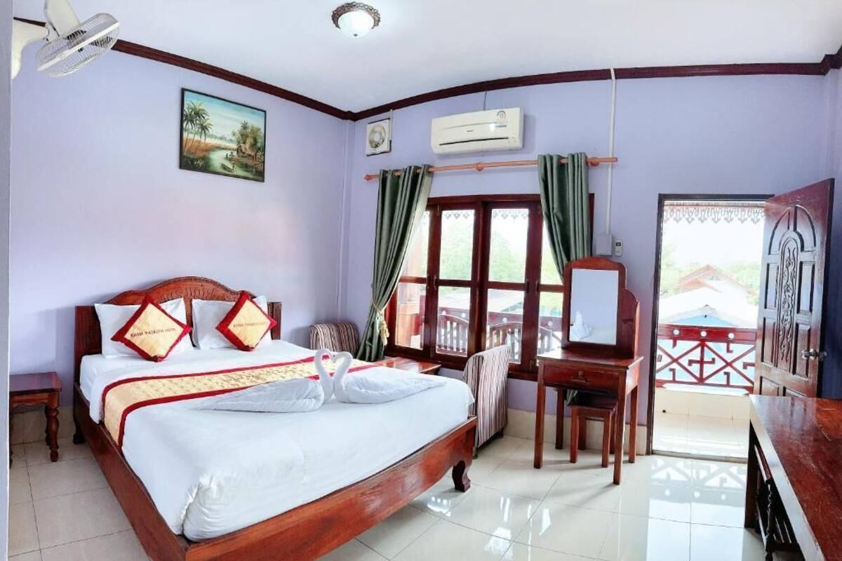 A hotel room with a bed, a desk, an air conditioner, a fan, a window with curtains, and a view of the outside.
