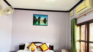 A hotel room with a bed, a painting of a landscape, a window with curtains, and an air conditioning unit. - Khampaseuth Hotel (Vang Vieng)