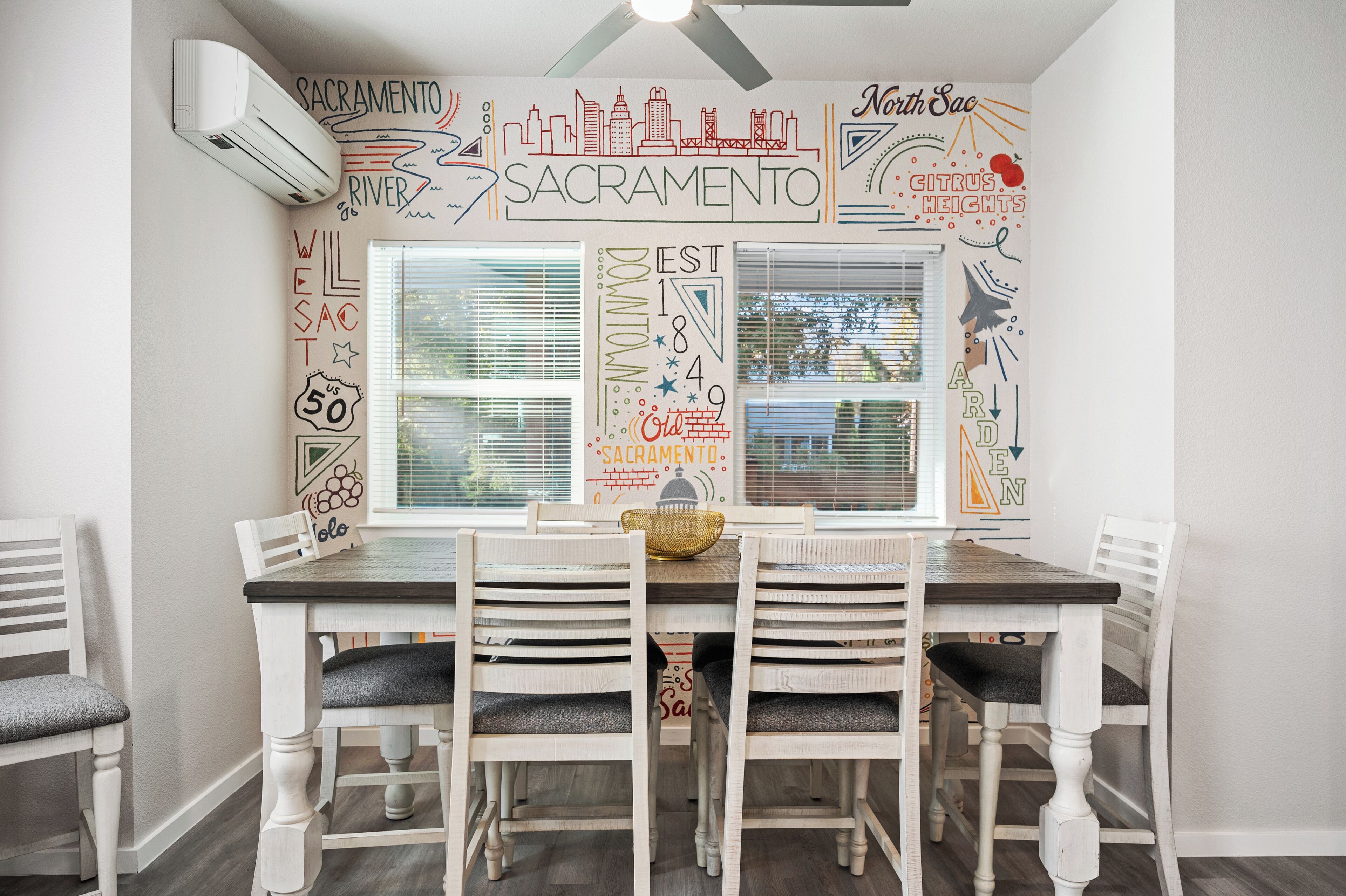 Un espace repas avec une table et des chaises blanches, une fresque murale sur le thème de la ville et une fenêtre avec des stores.