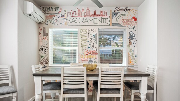 Un espace repas avec une table et des chaises blanches, une fresque murale sur le thème de la ville et une fenêtre avec des stores.