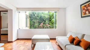 A modern living room with a large window, a grey sofa with orange cushions, a white ottoman, and a wooden herringbone floor. - LOFT  1-bedroom apartment in San Borja with WiFi (San Borja)