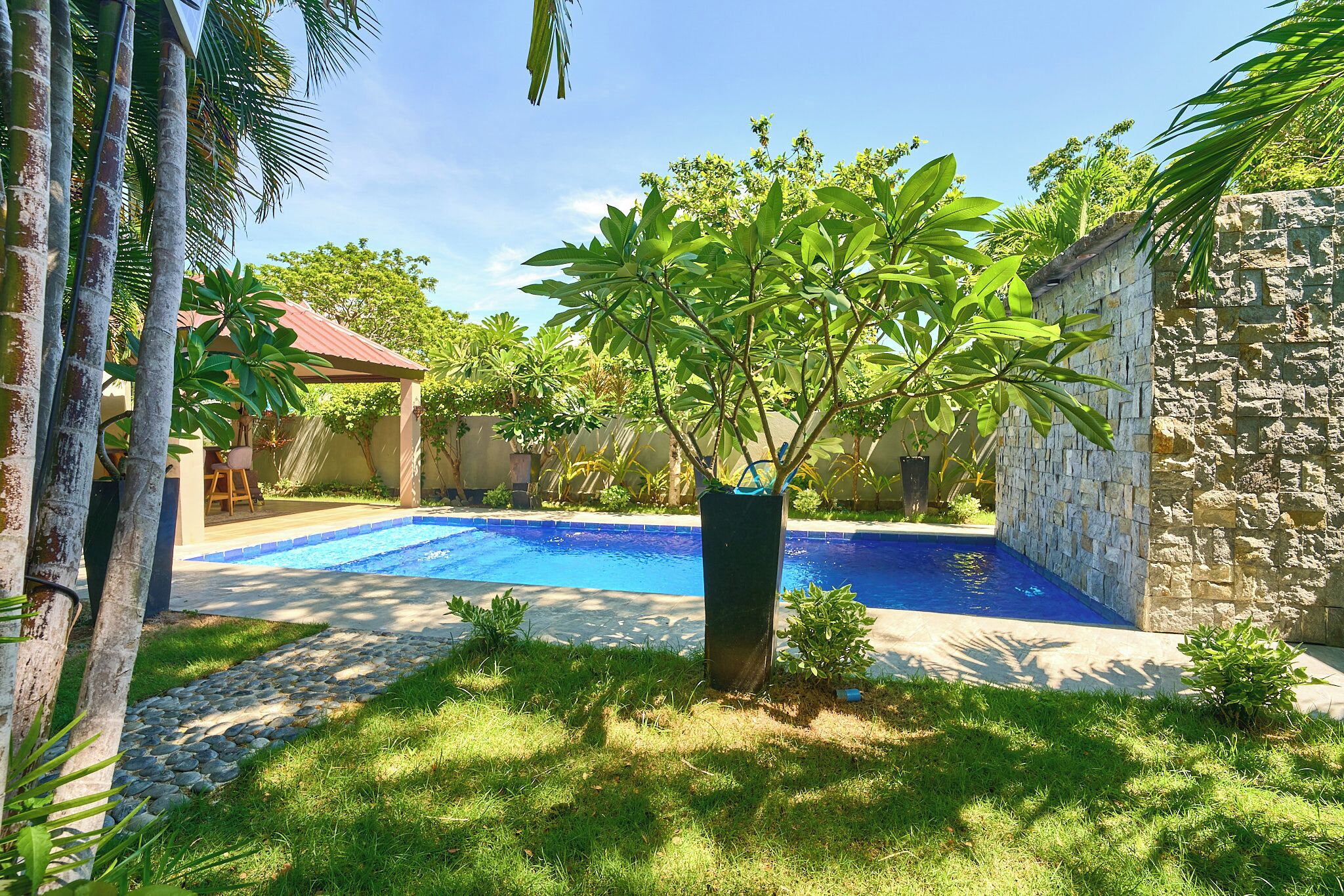 A swimming pool surrounded by a stone wall and lush greenery, with a small building in the background.
