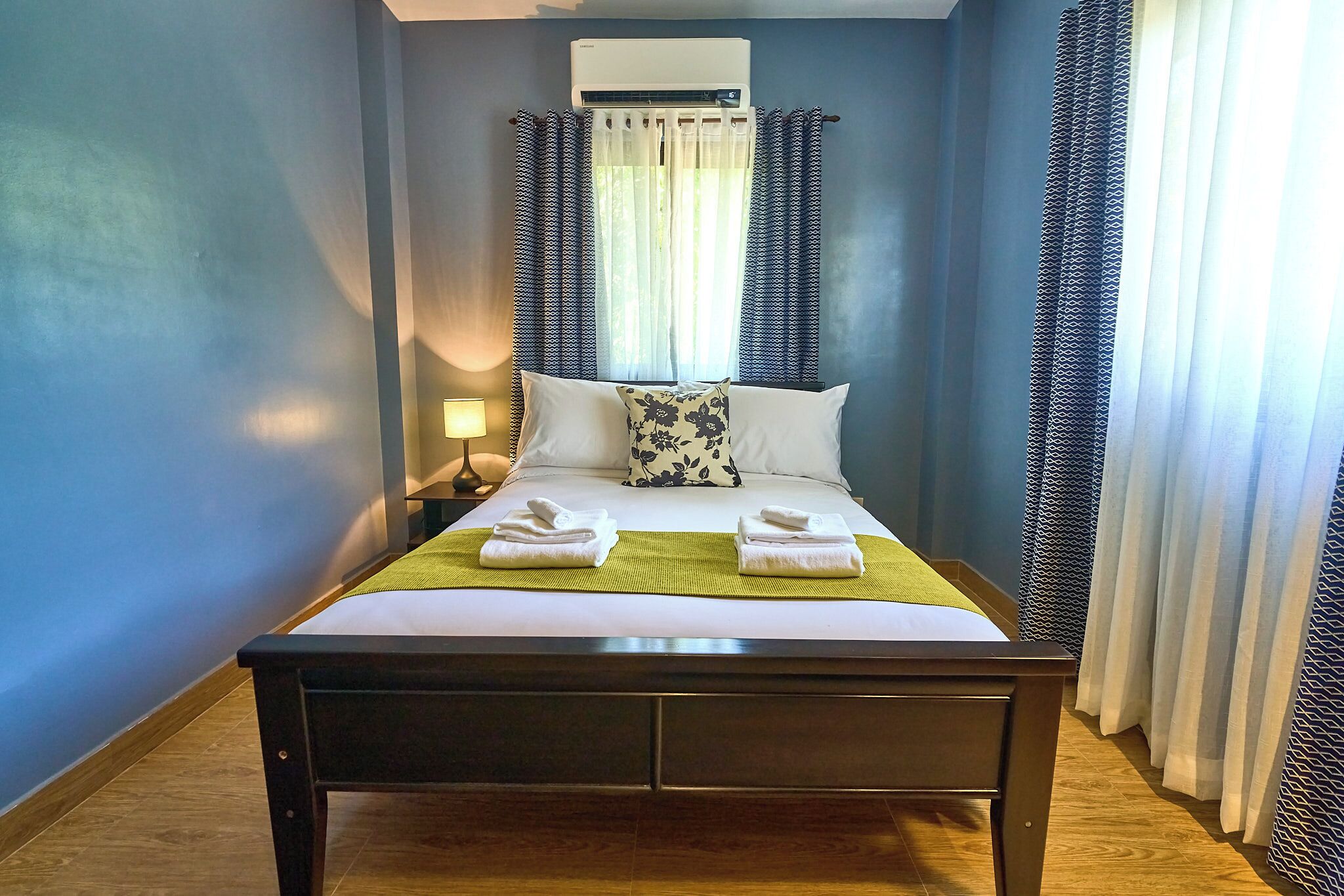 A neatly made bed with white linens and a green throw, a bedside lamp, and a window with patterned curtains.