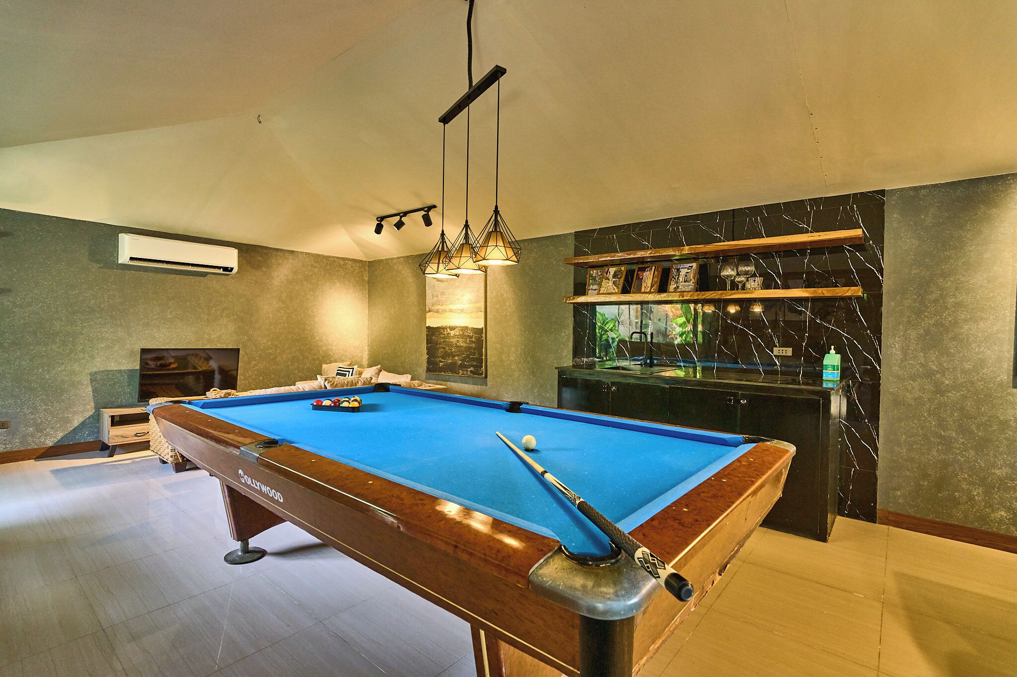 Une table de billard avec une queue de billard, une bille et un porte-queues. On y trouve également une étagère avec des objets décoratifs et un climatiseur fixé au mur.