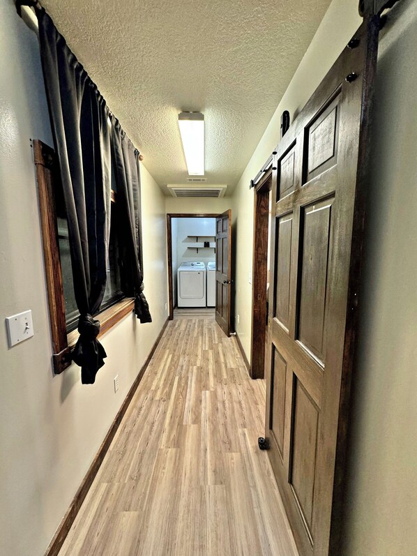 A hallway with wooden flooring, a ceiling vent, a door with a handle, and a window with curtains.
