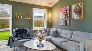 Un salon moderne avec un canapé gris, une table basse ronde et une grande fenêtre. On y trouve deux étagères murales ornées d’objets décoratifs et deux œuvres d’art encadrées.
