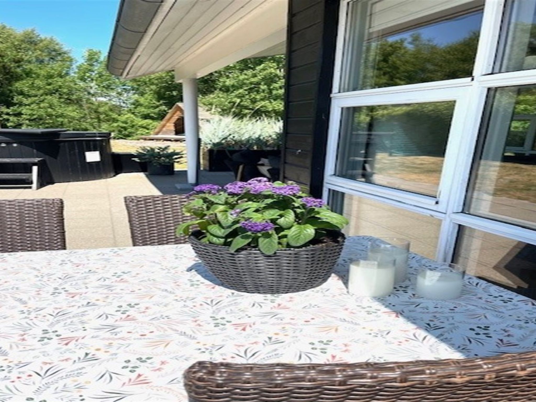 Un espace patio comprenant une table, des chaises en osier, un pot de fleurs avec des fleurs violettes et une fenêtre en verre.
