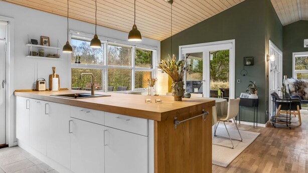Une cuisine moderne avec un îlot central en bois, des armoires blanches, des luminaires suspendus et une vue sur l’extérieur.