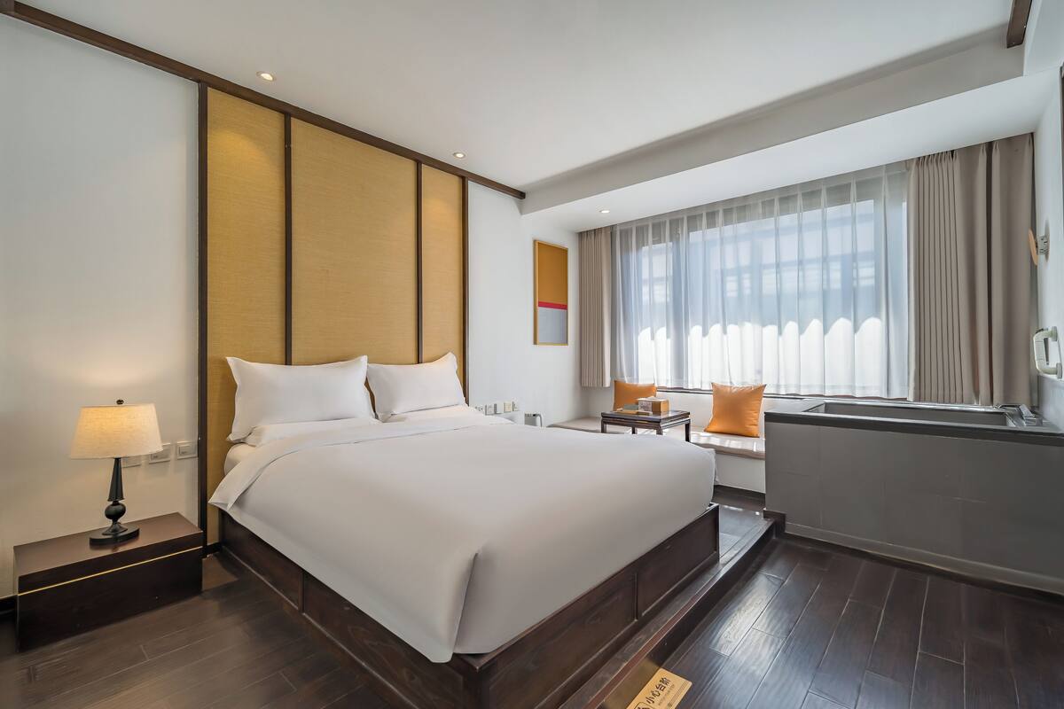 Une chambre d’hôtel avec un grand lit, une table de chevet avec une lampe, une petite table avec une chaise, une fenêtre avec des rideaux et une vue sur l’extérieur.