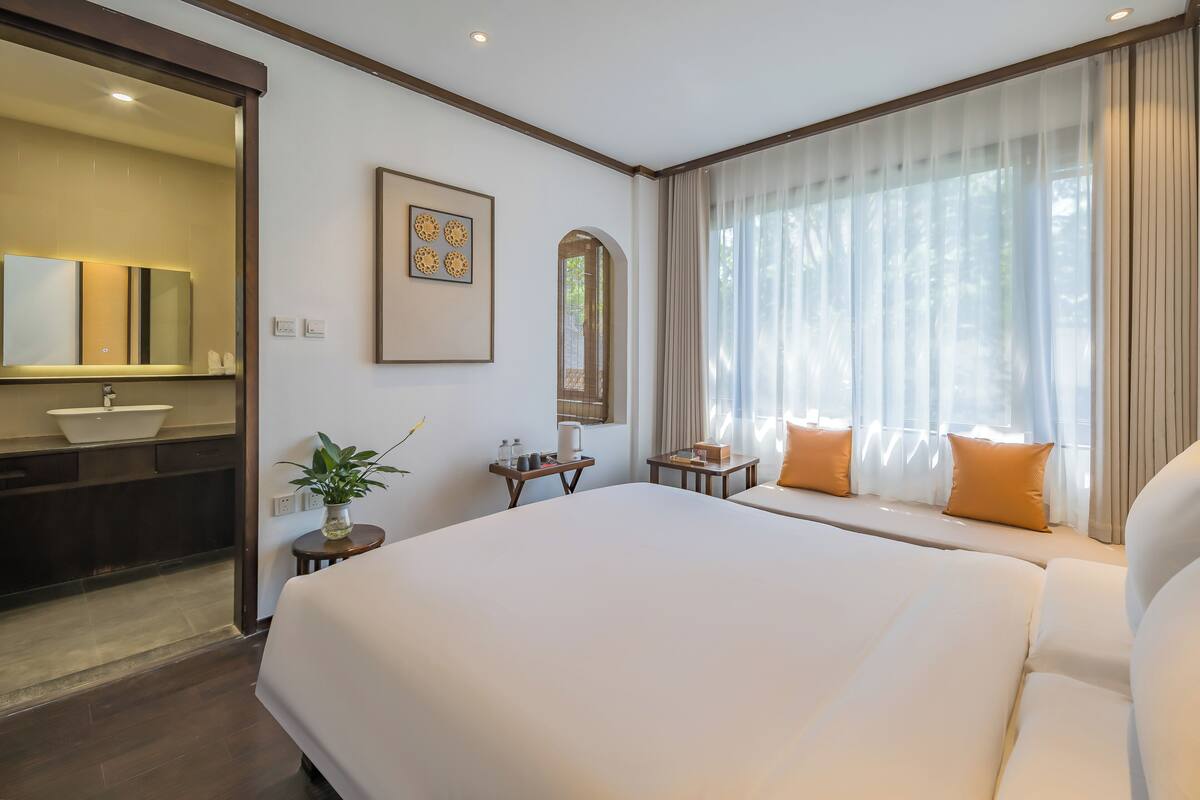 Une chambre d’hôtel avec un grand lit, une salle de bain avec un lavabo et un miroir, une petite table avec une plante et une fenêtre avec des rideaux.