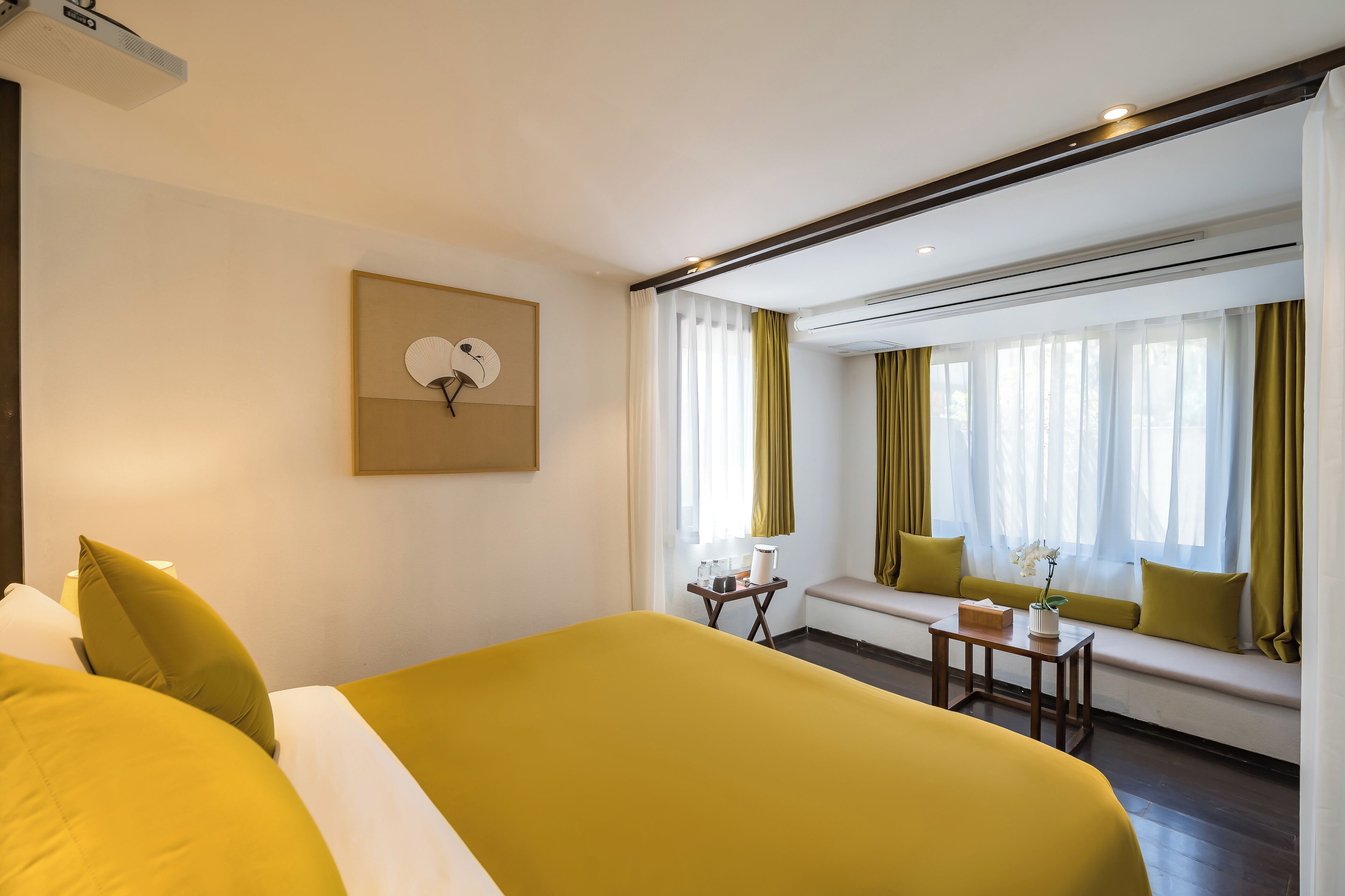 Ein Hotelzimmer mit einem großen Bett, einem kleinen Tisch mit einer Vase, einer Bank mit Kissen und einem Wandventilator.