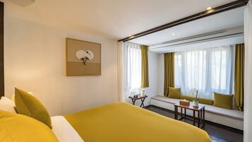 Ein Hotelzimmer mit einem großen Bett, einem kleinen Tisch mit einer Vase, einer Bank mit Kissen und einem Wandventilator.