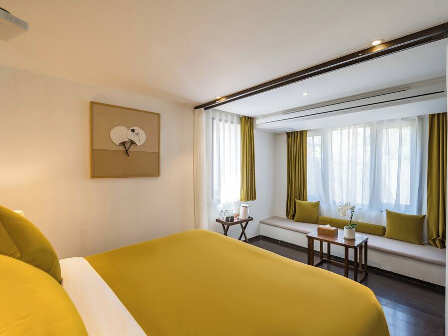 Ein Hotelzimmer mit einem großen Bett, einem kleinen Tisch mit einer Vase, einer Bank mit Kissen und einem Wandventilator.