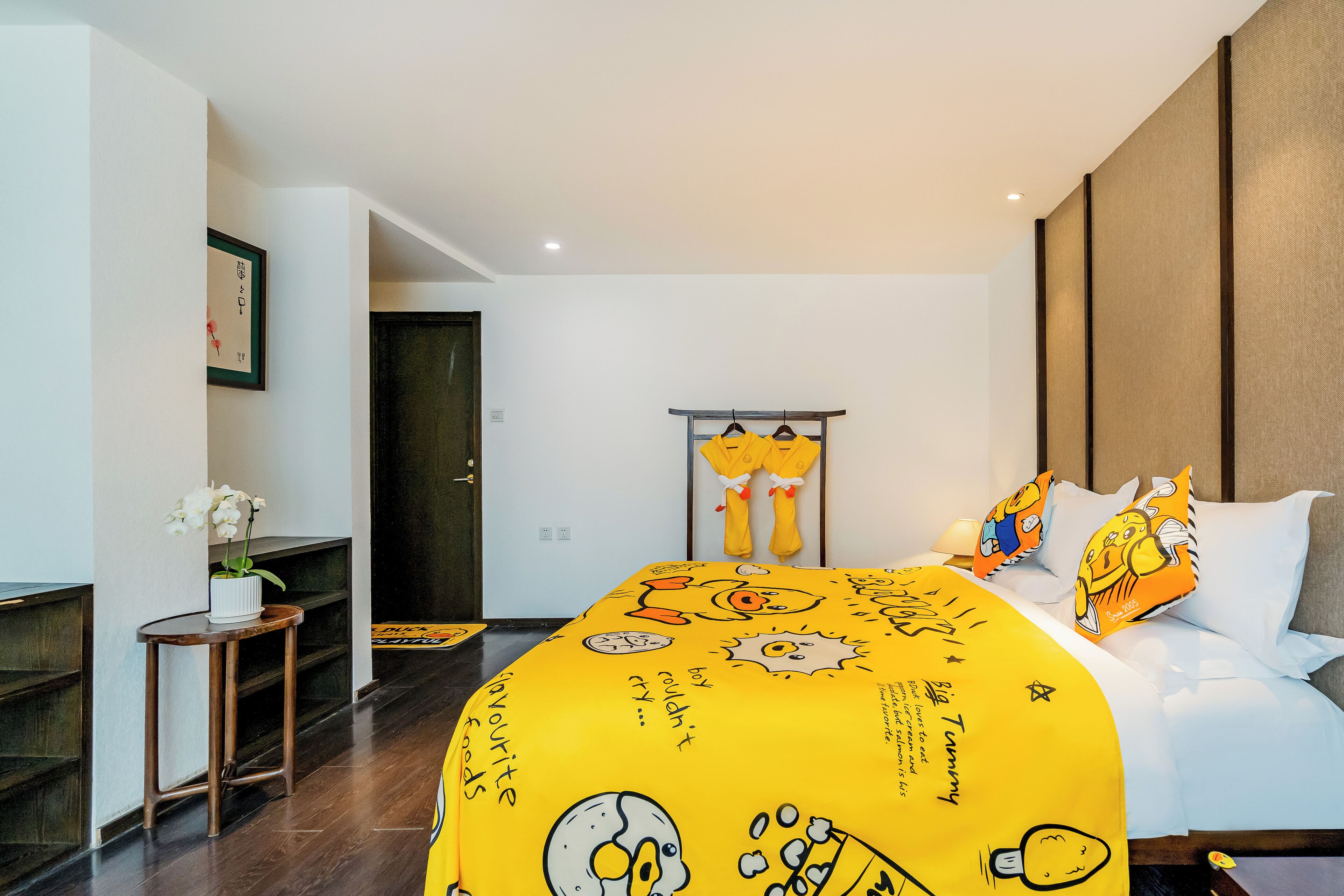 Quarto de hotel moderno com colcha amarela vibrante com personagens de desenho animado, uma pequena mesa de cabeceira com um vaso de flores e piso de madeira.