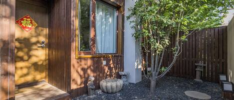 A wooden house entrance with a decorative door, a window, and a small garden with stepping stones and a tree.