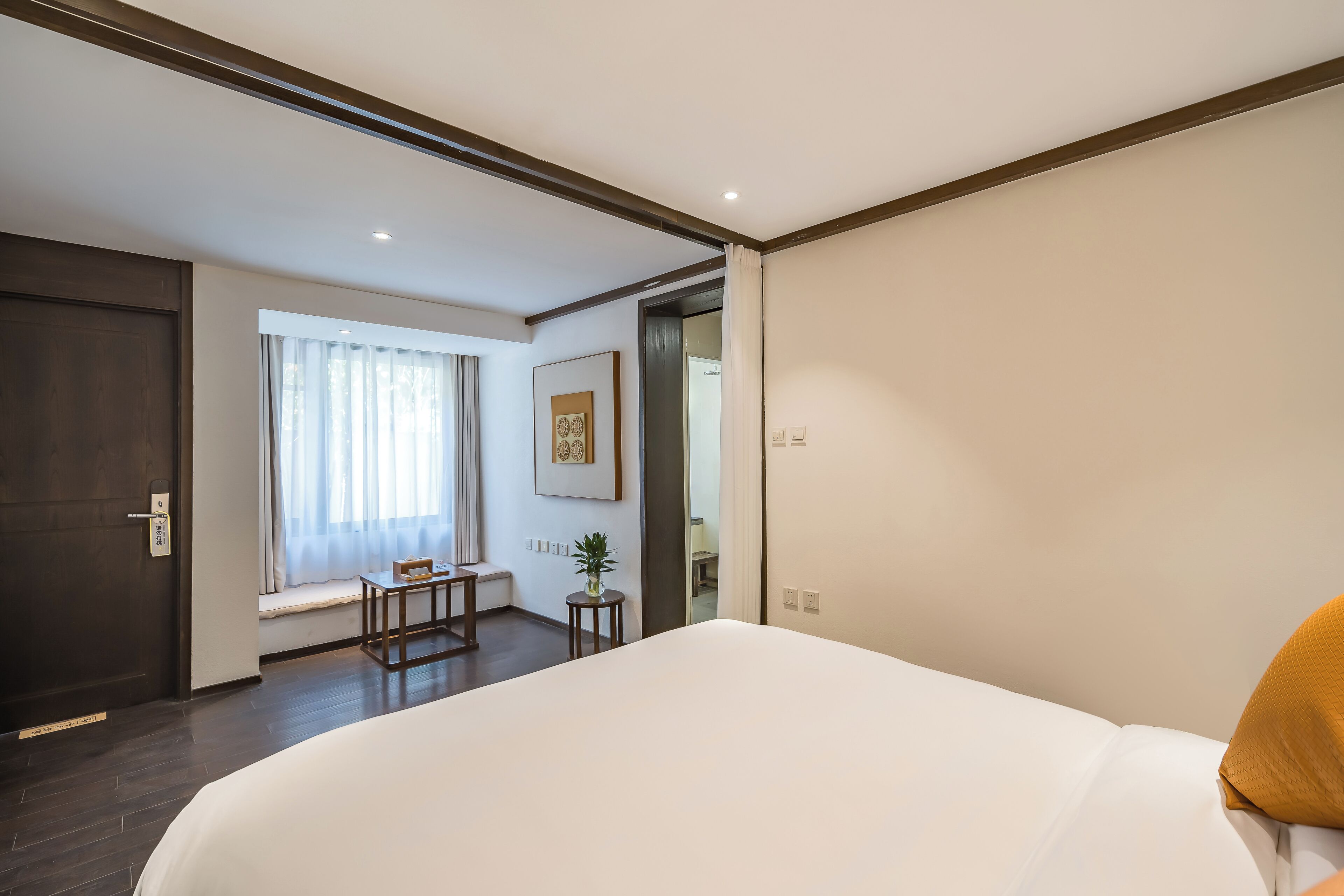 Una habitación de hotel con una cama grande, una mesita pequeña, una silla, una ventana con cortinas, un cuadro enmarcado en la pared y una puerta.