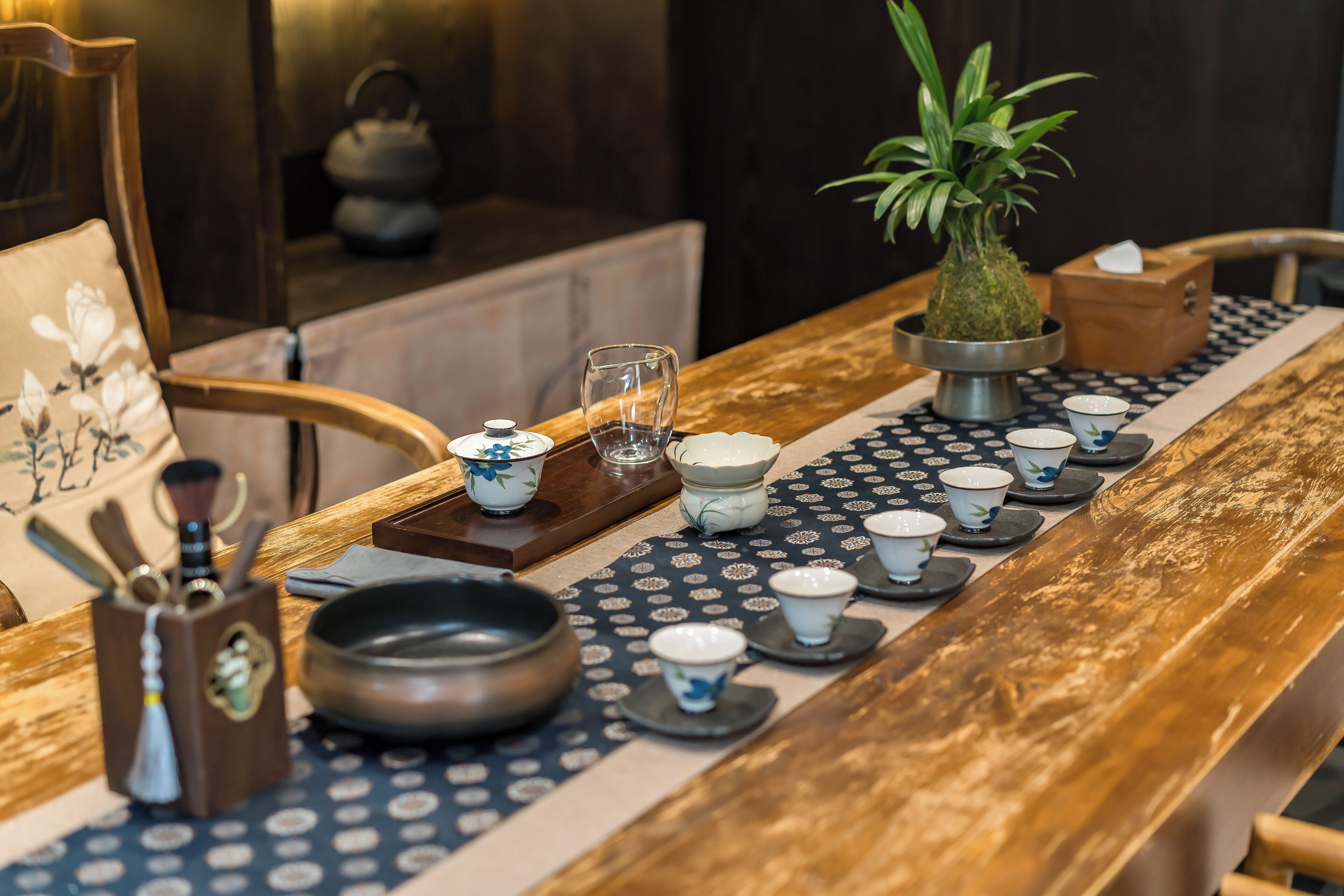 A wooden dining table with a patterned table runner, a potted plant, and a tea set.