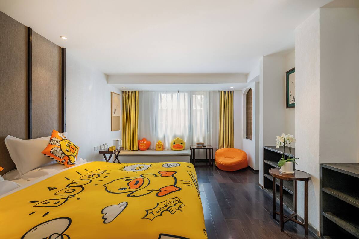 Une chambre d’hôtel avec un lit orné d’un motif représentant un personnage de dessin animé, une fenêtre avec des rideaux, une petite table avec un vase et une chaise.