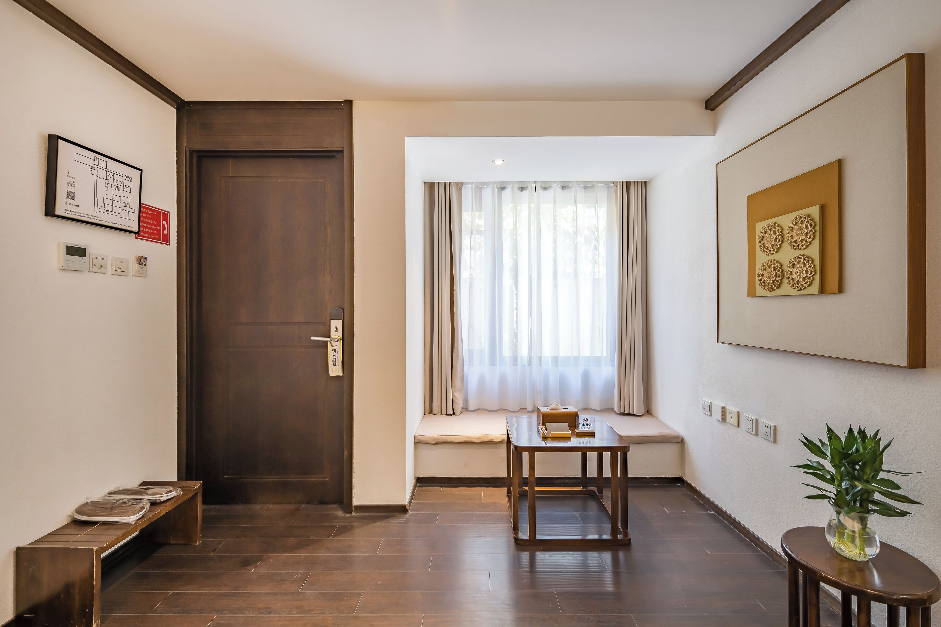 A hotel room with a wooden door, a bench, a table, a window with curtains, and a wall-mounted artwork.