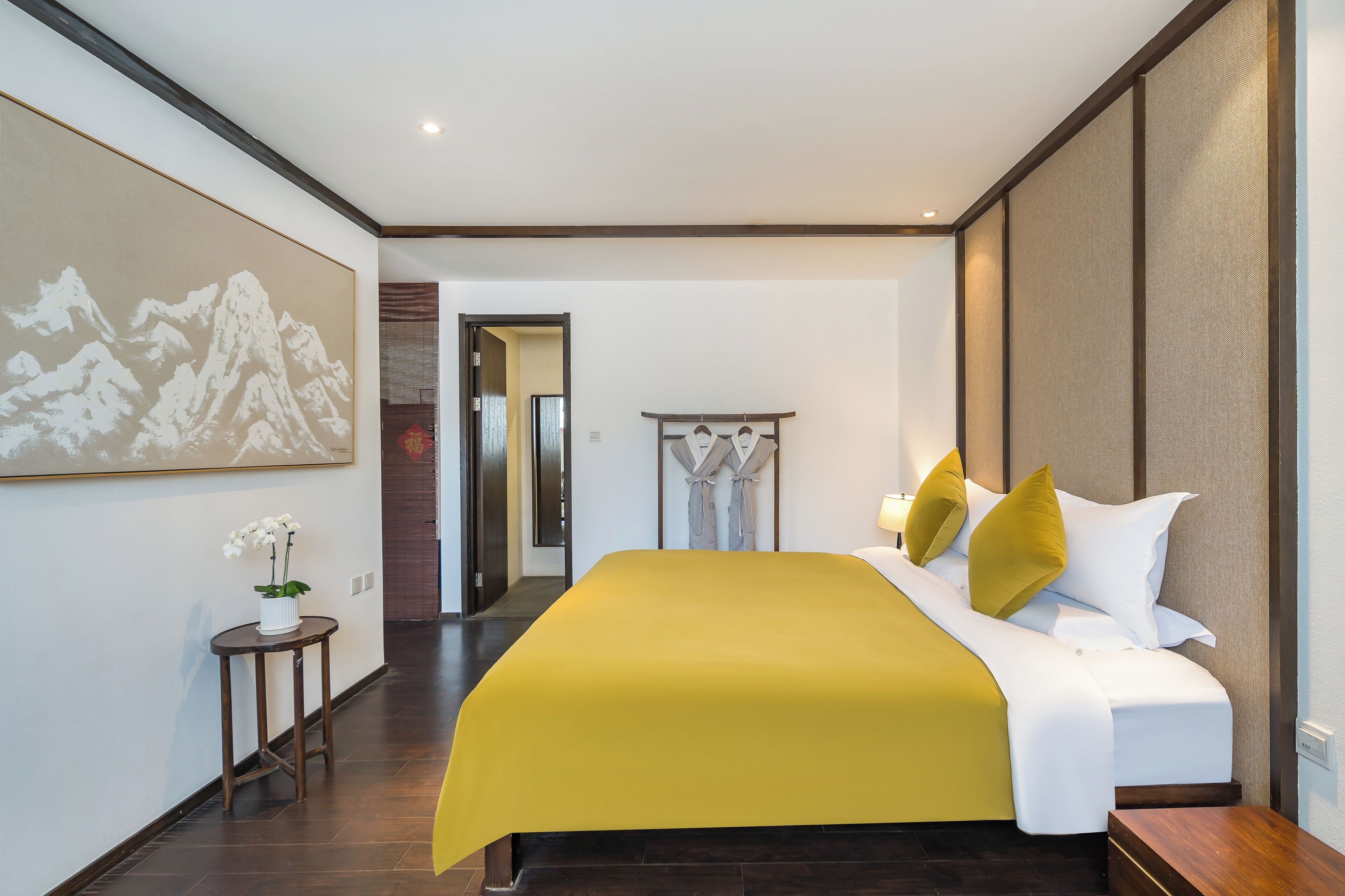 A hotel room with a large bed, a bedside table, a small table with a plant, a painting on the wall, and a hallway leading to another room.