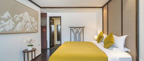 Ein Hotelzimmer mit einem großen Bett, einem Nachttisch, einem kleinen Tisch mit einer Pflanze, einem Bild an der Wand und einem Flur, der in ein weiteres Zimmer führt.