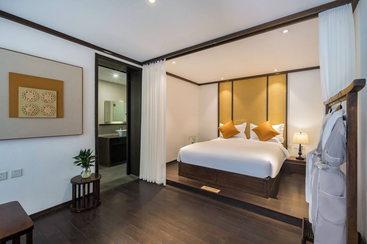 Une chambre d’hôtel moderne avec un grand lit, une table de chevet avec une lampe, un peignoir suspendu à un porte-vêtements en bois et une petite plante en pot.
