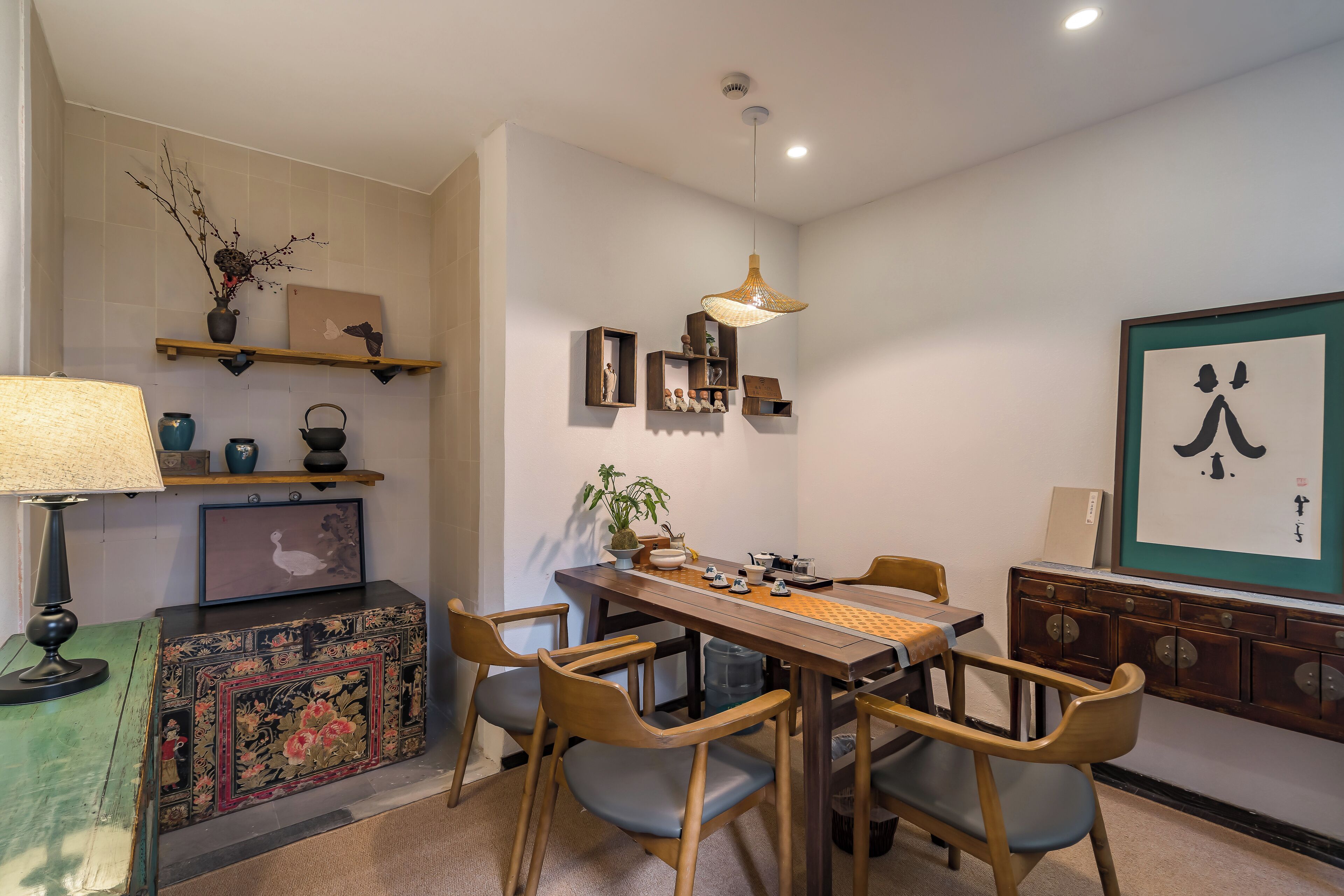 A dining area with a wooden table, chairs, a hanging light fixture, and a wall-mounted shelf with decorative items.