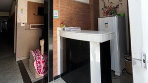 Zona de recepción de un hotel, con mostrador, candelabro decorativo, una planta pequeña y un refrigerador.
