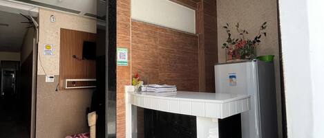 Zona de recepción de un hotel, con mostrador, candelabro decorativo, una planta pequeña y un refrigerador.