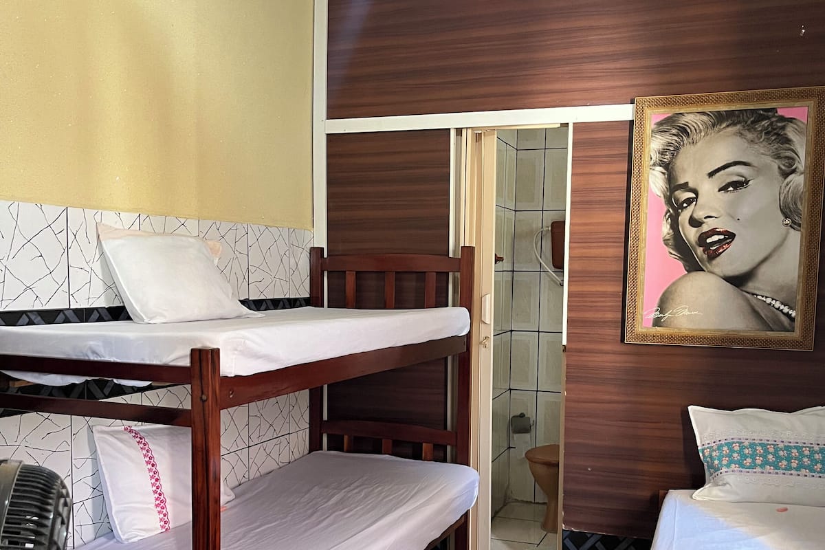 Une chambre avec un lit superposé, un lit simple, un ventilateur de plafond, une salle de bain avec des toilettes et un tableau encadré au mur.