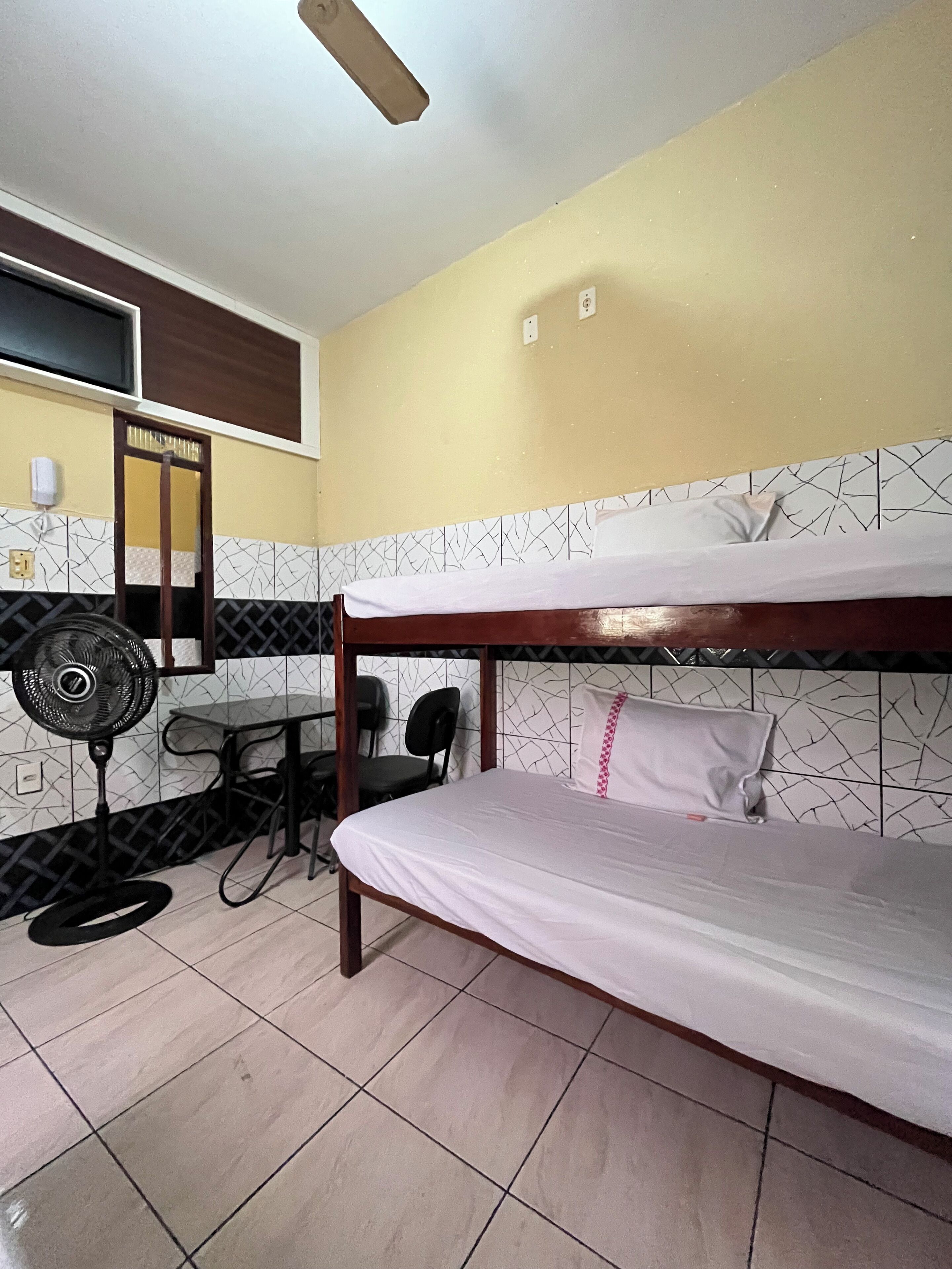 A room with a bunk bed, a desk with a chair, a standing fan, and a tiled wall with a geometric pattern.